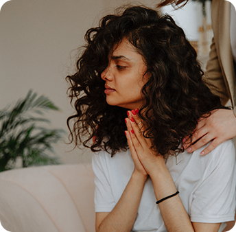 A person with curly hair sitting pensively with hands clasped, while another person's hands rest on their shoulder.